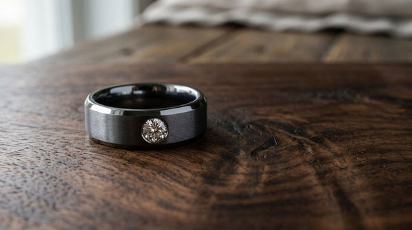 A luxury men's tungsten wedding band featuring a round diamond inlay resting on a wooden table.